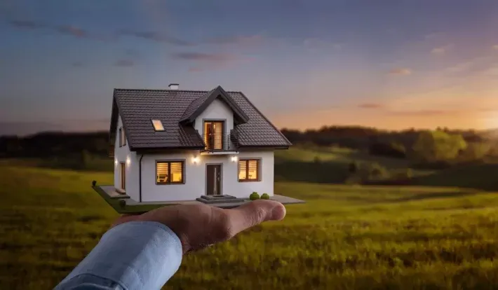 Mão segurando modelo de uma casa com um terreno rural ao fundo.